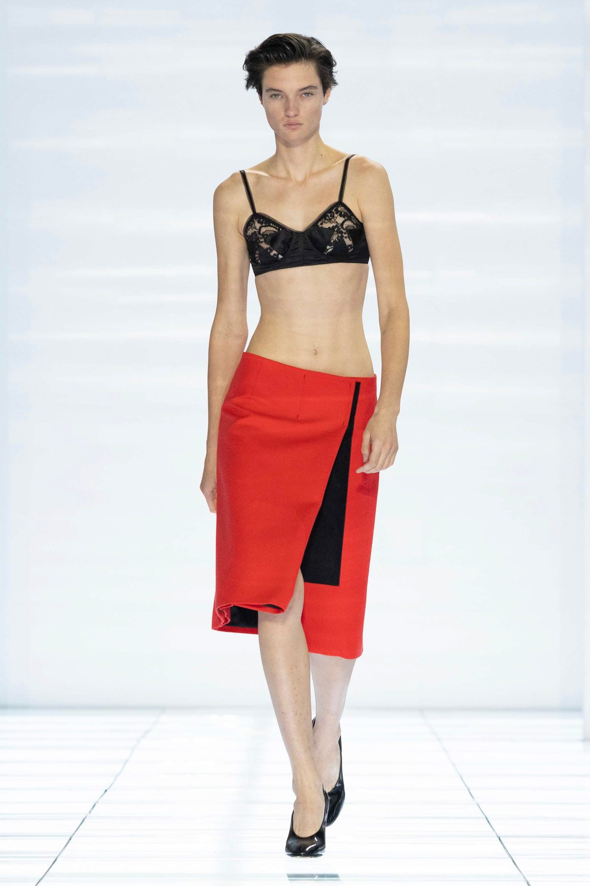 Model wearing a red asymmetric Prada skirt and black lace bra on the Spring 2027 runway
