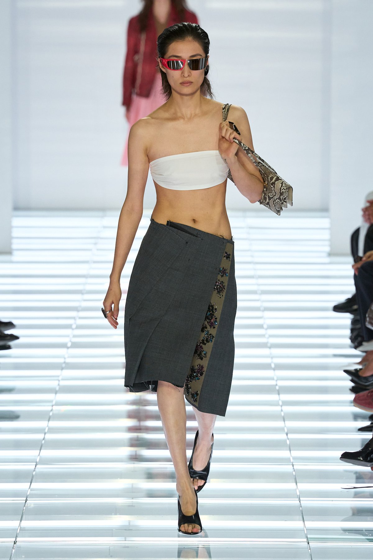 Prada Spring 2027 look with a grey asymmetric skirt, white bandeau, pink shield sunglasses and snakeskin shoulder bag