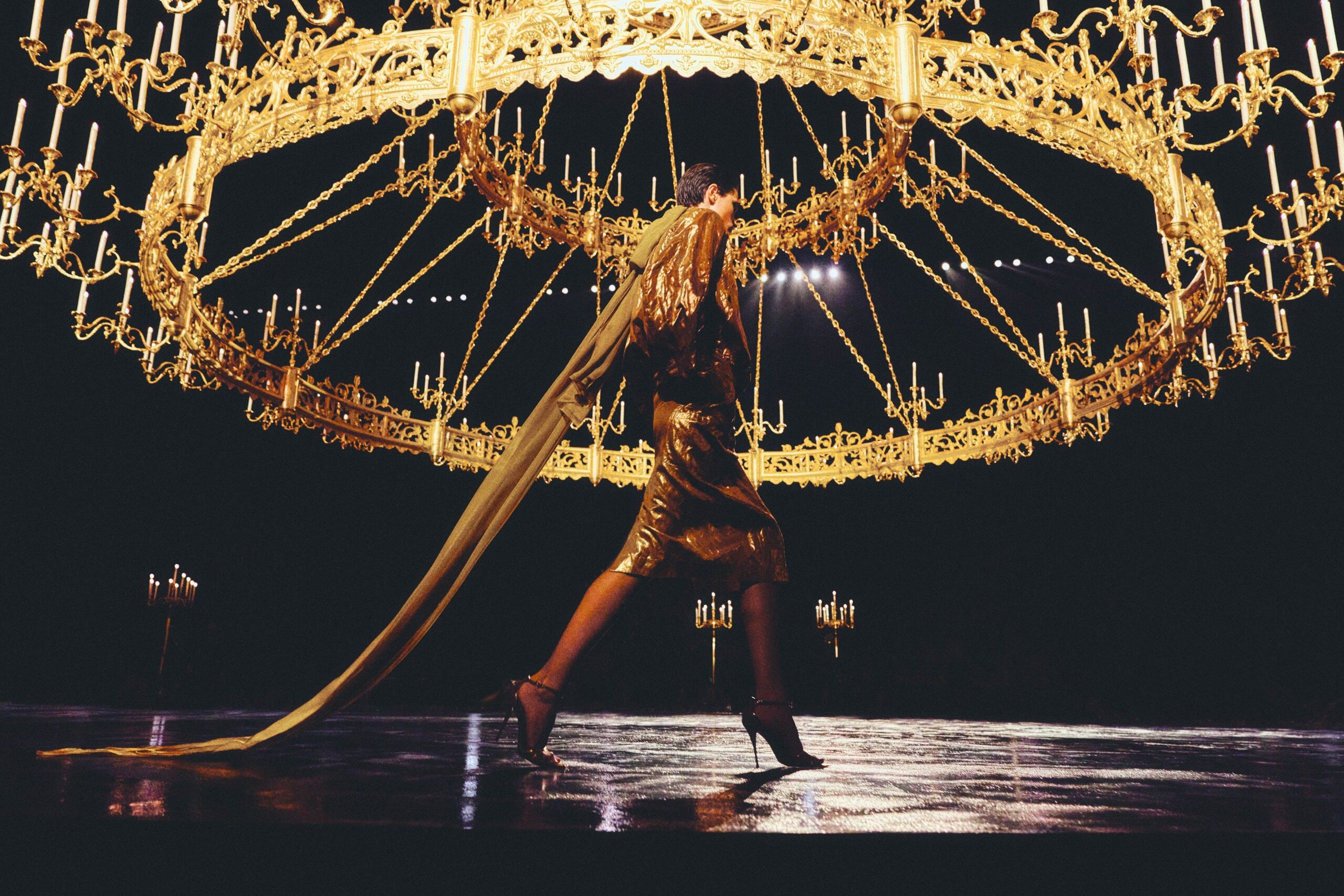 Saint Laurent Summer 2027 gold look walking beneath an enormous chandelier